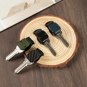 May include: Four compact key organizers with metal keys attached. The organizers feature different top plates: one olive green, one solid black, one dark blue, and one black with a carbon fiber pattern. Each unit has a folding mechanism.
