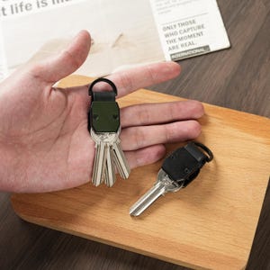 May include: Two key organizers featuring a metallic top plate and a black leather strap with a circular ring. One organizer is olive green and holds three silver keys, while the other is black and holds a single silver key with a small engraved logo.