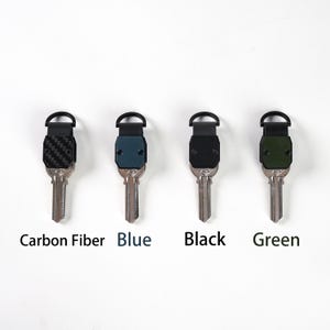 May include: Four key organizers with metal key blades, each featuring a distinct top plate. The plates are labeled Carbon Fiber, Blue, Black, and Green. Each organizer includes a black semi-circular attachment loop at the top for keychains.