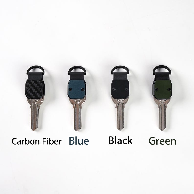 May include: Four key organizers with metal key blades, each featuring a distinct top plate. The plates are labeled Carbon Fiber, Blue, Black, and Green. Each organizer includes a black semi-circular attachment loop at the top for keychains.