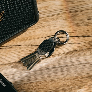 May include: A metal keychain featuring a black carbon fiber textured plate attached to a circular ring. The keychain holds a set of silver-colored keys, with the hardware displaying a dark metallic finish and a compact, structured design.