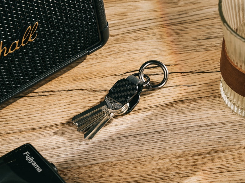 May include: A metal keychain featuring a black carbon fiber textured plate attached to a circular ring. The keychain holds a set of silver-colored keys, with the hardware displaying a dark metallic finish and a compact, structured design.