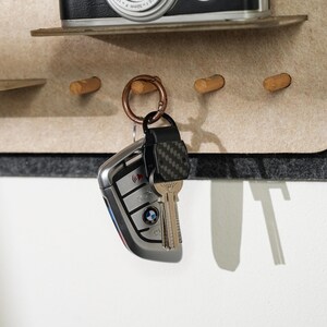 May include: A set of keys with a silver BMW car fob and a metallic key attached to a dark carbon fiber patterned fob, all suspended from a copper-colored circular ring hanging on a wooden peg mounted to a beige felt wall organizer board.