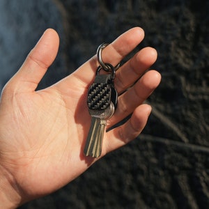May include: A metal keychain featuring a black carbon fiber textured square plate attached to a silver-toned split ring. The keychain holds several standard silver-colored metal keys that hang vertically from the central attachment point.