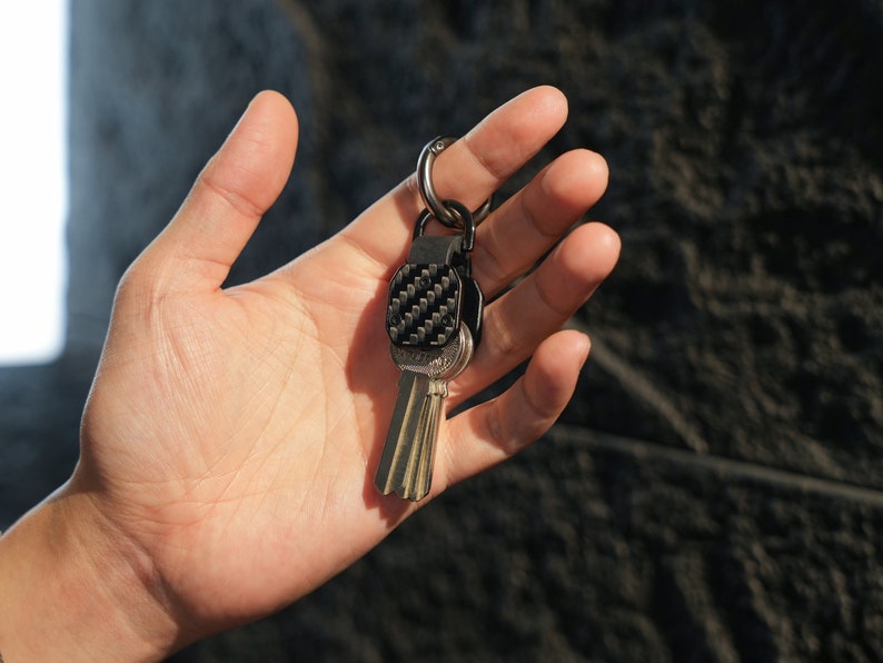 May include: A metal keychain featuring a black carbon fiber textured square plate attached to a silver-toned split ring. The keychain holds several standard silver-colored metal keys that hang vertically from the central attachment point.
