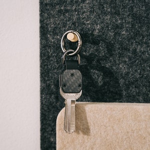 May include: A key organizer featuring a black carbon fiber textured plate with a metal key attached. The assembly includes a dark metal ring and a black leather strap, suspended from a small wooden peg. The key has a metallic finish.