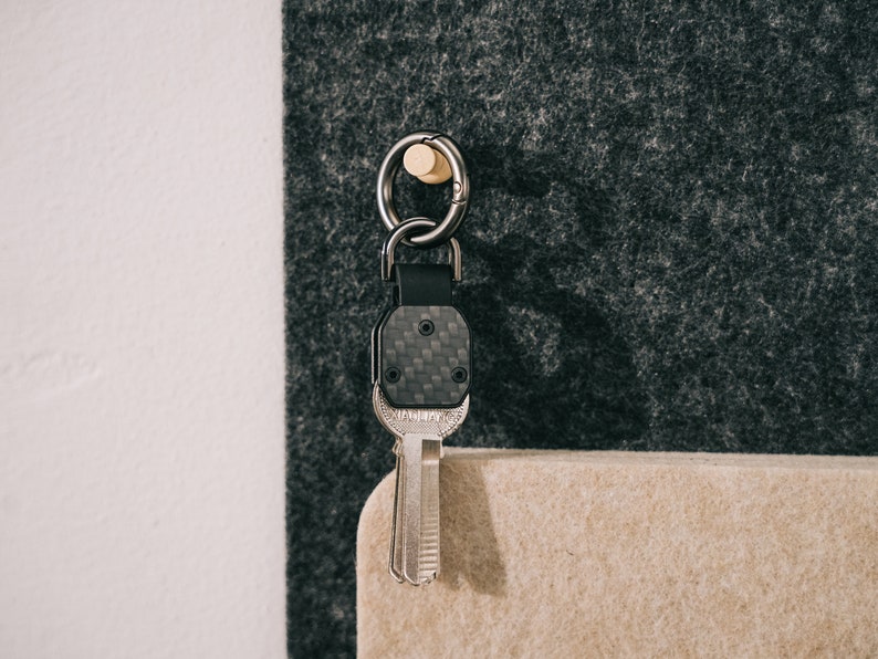 May include: A key organizer featuring a black carbon fiber textured plate with a metal key attached. The assembly includes a dark metal ring and a black leather strap, suspended from a small wooden peg. The key has a metallic finish.