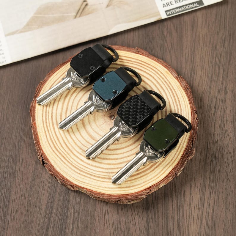 May include: Four metal keys attached to individual folding organizers. The organizers feature different finishes: matte black, metallic blue, black carbon fiber pattern, and olive green. Each organizer has a small black metal ring for attachment.
