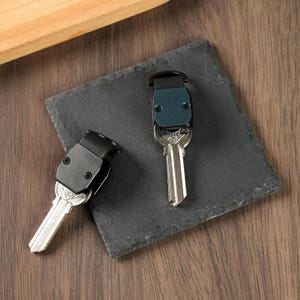 May include: Two key organizers with metallic silver keys attached. One organizer features a matte black casing, while the other has a dark blue casing. Both organizers have a rectangular shape with two visible screws on the front face.
