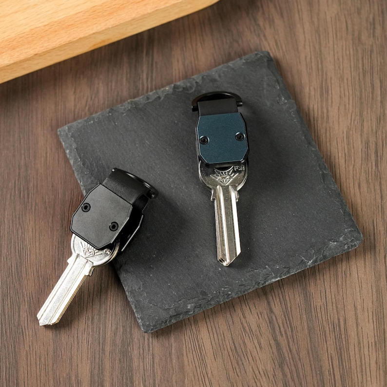 May include: Two key organizers with metallic silver keys attached. One organizer features a matte black casing, while the other has a dark blue casing. Both organizers have a rectangular shape with two visible screws on the front face.