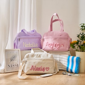 May include: Three pastel-coloured baby bags, in lavender, pink, and cream, each embroidered with a name. The bags have top handles, shoulder straps, and front pockets. A framed print and swimming accessories are also visible in the image.