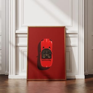Puede incluir: Un cuadro enmarcado con una vista superior de un coche deportivo rojo sobre un fondo rojo. El coche tiene un interior negro y está enmarcado en madera. El cuadro se exhibe sobre un suelo de madera.
