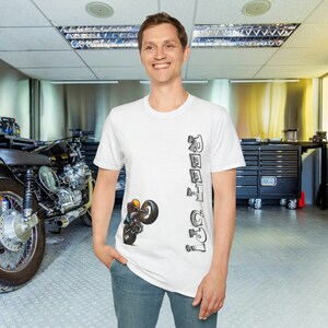 Peut inclure: T-shirt blanc avec un motif de moto et le texte "REET UP!". Le motif représente une moto faisant une roue arrière. Le t-shirt est à col rond et est porté par une personne.