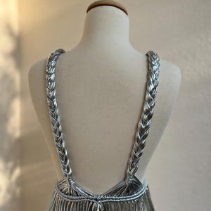 May include: A silver dress displayed on a mannequin. The dress features braided straps and a fringe-like skirt. The metallic material reflects light, creating a shimmering effect. The dress is designed with a low back and a unique, eye-catching style.