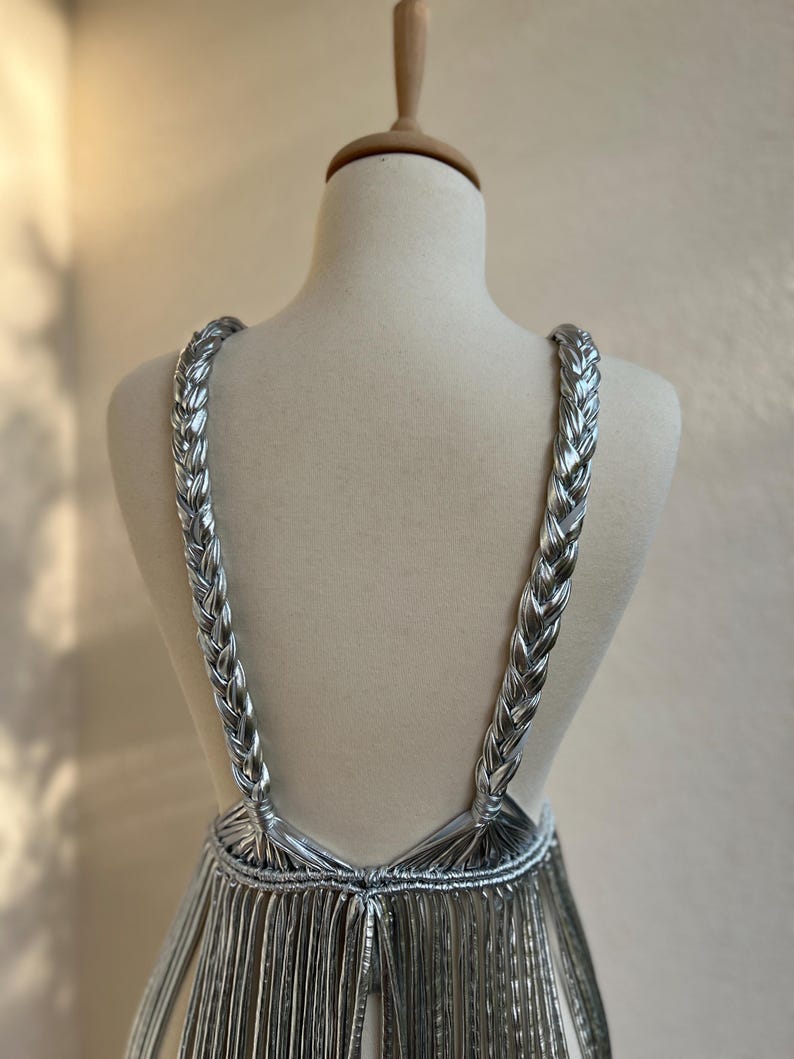 May include: A silver dress displayed on a mannequin. The dress features braided straps and a fringe-like skirt. The metallic material reflects light, creating a shimmering effect. The dress is designed with a low back and a unique, eye-catching style.
