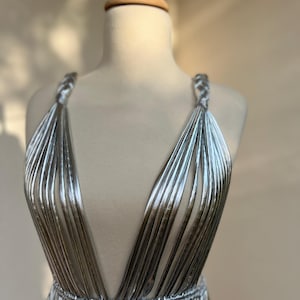 May include: A silver, sleeveless dress with a deep V-neck and a cinched waist. The dress is made of vertical, pleated fabric that creates a flowing, elegant look. The straps are braided, and the dress is displayed on a mannequin.
