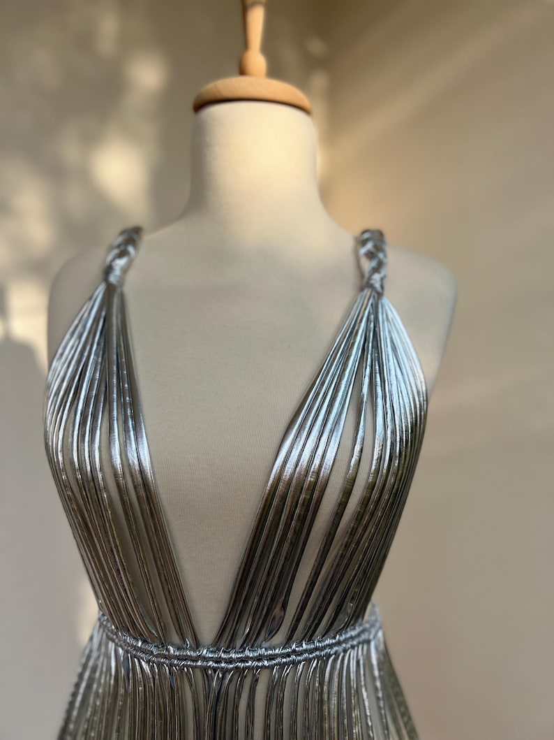 May include: A silver, sleeveless dress with a deep V-neck and a cinched waist. The dress is made of vertical, pleated fabric that creates a flowing, elegant look. The straps are braided, and the dress is displayed on a mannequin.