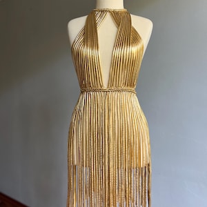 Puede incluir: Vestido dorado de estilo halter con escote en V profundo y flecos. El vestido se exhibe en un maniqu&iacute;. El vestido est&aacute; hecho de material dorado texturizado vertical y tiene un cintur&oacute;n tipo cuerda alrededor de la cintura. El vestido mide unos 80 cm de largo.