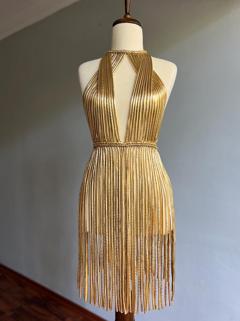 Puede incluir: Vestido dorado de estilo halter con escote en V profundo y flecos. El vestido se exhibe en un maniqu&iacute;. El vestido est&aacute; hecho de material dorado texturizado vertical y tiene un cintur&oacute;n tipo cuerda alrededor de la cintura. El vestido mide unos 80 cm de largo.