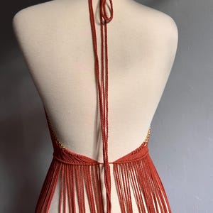 May include: A terracotta-colored halter top with a fringe bottom. The top is made of a crocheted material and has a thin gold chain detail.