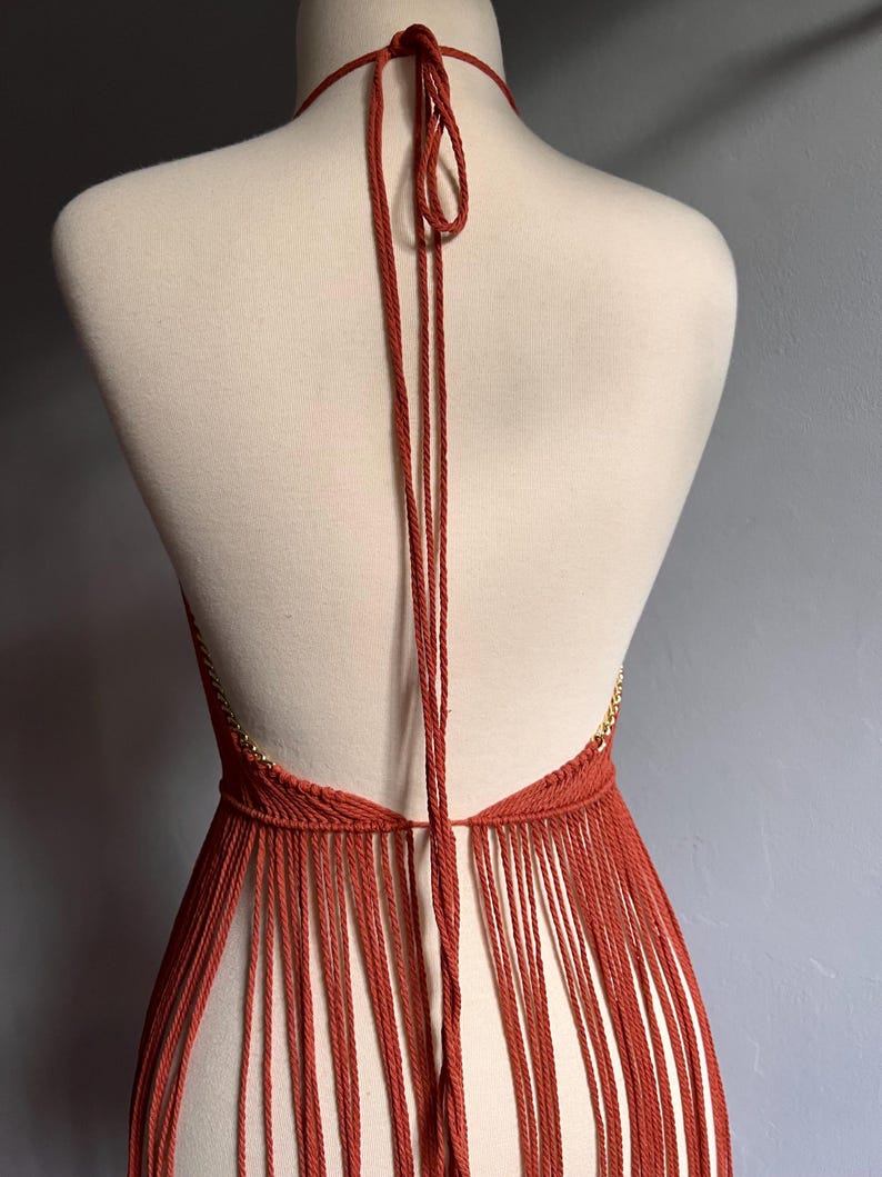 May include: A terracotta-colored halter top with a fringe bottom. The top is made of a crocheted material and has a thin gold chain detail.