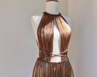 May include: A copper-colored, handmade dress displayed on a mannequin. The dress features a halter neckline, a cut-out at the chest, and a fringed skirt. The dress is made of vertical, thin copper rods, and has a wrapped waist.