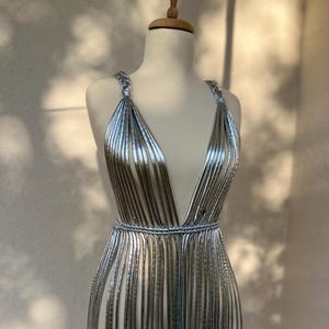 May include: A silver, sleeveless dress with a plunging neckline and a fringed skirt. The dress is displayed on a mannequin. The dress has a metallic sheen and a cinched waist. The dress is made of vertical strips of silver material.