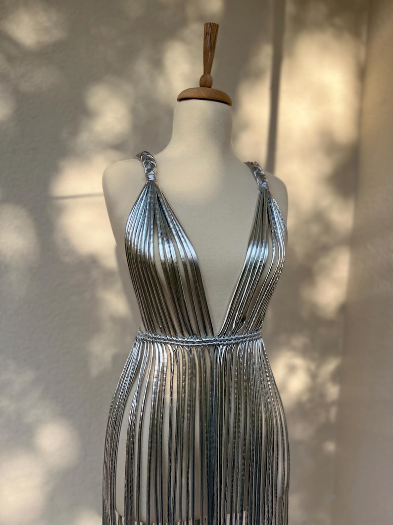 May include: A silver, sleeveless dress with a plunging neckline and a fringed skirt. The dress is displayed on a mannequin. The dress has a metallic sheen and a cinched waist. The dress is made of vertical strips of silver material.