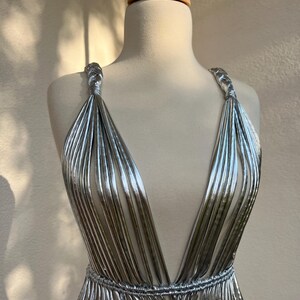 May include: A silver, pleated dress with a plunging neckline and braided straps. The dress is displayed on a mannequin, showcasing its flowing, metallic design. The waist is cinched with a matching pleated band.