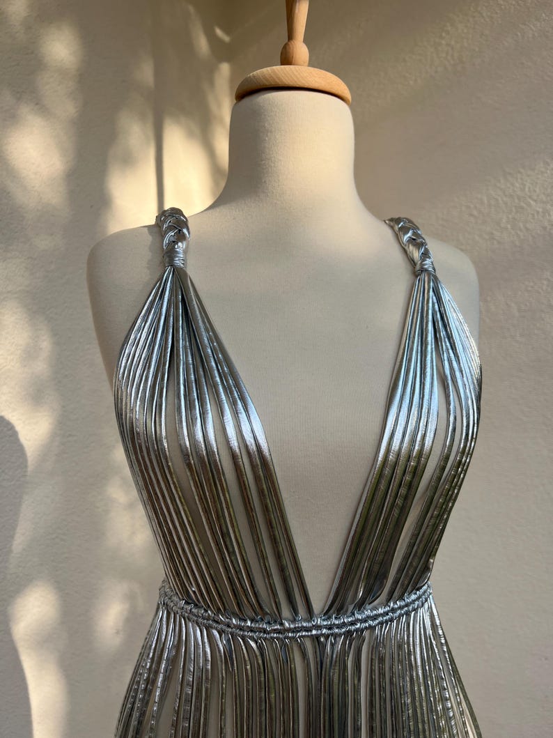 May include: A silver, pleated dress with a plunging neckline and braided straps. The dress is displayed on a mannequin, showcasing its flowing, metallic design. The waist is cinched with a matching pleated band.