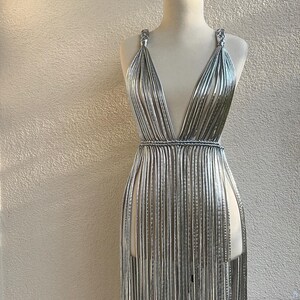 May include: A silver dress on a mannequin. The dress features a plunging neckline and is constructed from numerous vertical strands, creating a fringe effect. The dress is displayed on a wooden mannequin against a neutral wall.