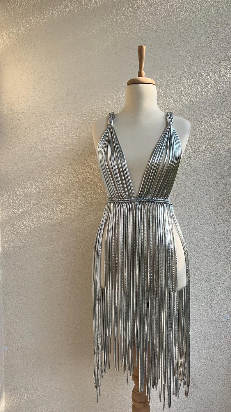 May include: A silver dress on a mannequin. The dress features a plunging neckline and is constructed from numerous vertical strands, creating a fringe effect. The dress is displayed on a wooden mannequin against a neutral wall.