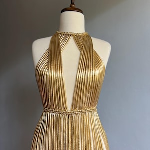 Puede incluir: Un vestido dorado de estilo halter con escote pronunciado. El vestido est&aacute; hecho de tela plisada vertical que crea un efecto fluido. El vestido se exhibe en un maniqu&iacute;.