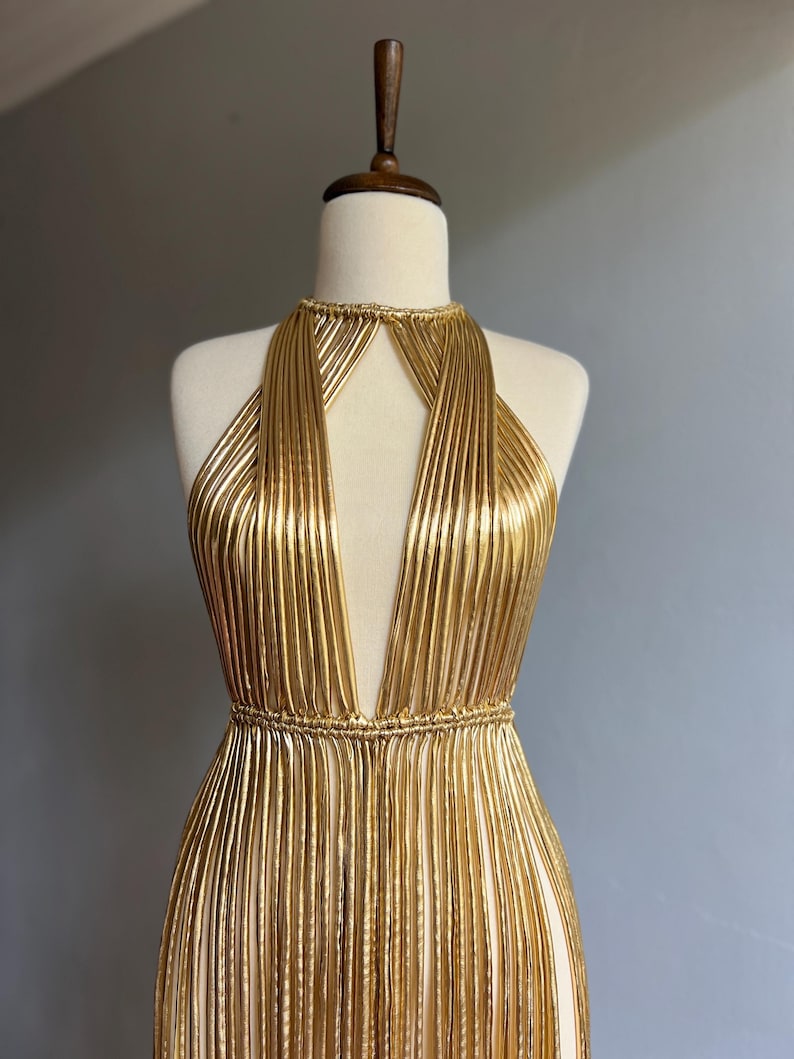 Puede incluir: Un vestido dorado de estilo halter con escote pronunciado. El vestido est&aacute; hecho de tela plisada vertical que crea un efecto fluido. El vestido se exhibe en un maniqu&iacute;.