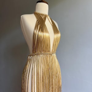 Puede incluir: Vestido dorado de estilo halter con escote pronunciado y detalles de flecos. El vestido se exhibe en un maniqu&iacute;, mostrando su intrincado dise&ntilde;o y silueta fluida. El vestido parece estar hecho de material met&aacute;lico.