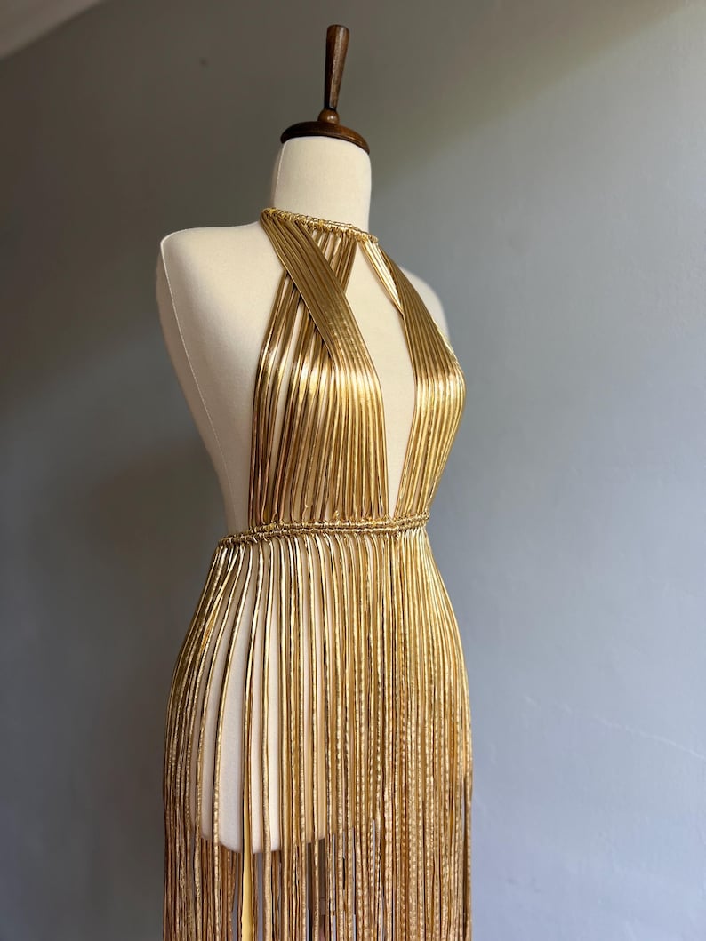 Puede incluir: Vestido dorado de estilo halter con escote pronunciado y detalles de flecos. El vestido se exhibe en un maniqu&iacute;, mostrando su intrincado dise&ntilde;o y silueta fluida. El vestido parece estar hecho de material met&aacute;lico.