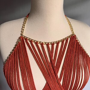 May include: A red crocheted halter top with a gold chain detail. The top is made of many thin strands of yarn that are woven together to create a criss-cross pattern.