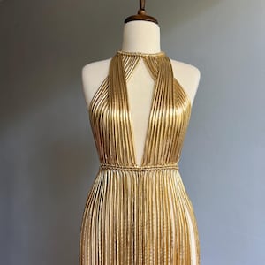 Puede incluir: Vestido dorado de estilo halter con escote pronunciado y falda con flecos, expuesto en un maniqu&iacute;. El vestido presenta hebras doradas verticales, creando una silueta fluida. Un llamativo ejemplo de ropa de noche.