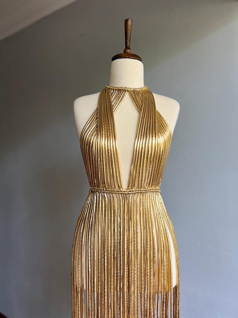 Puede incluir: Vestido dorado de estilo halter con escote pronunciado y falda con flecos, expuesto en un maniqu&iacute;. El vestido presenta hebras doradas verticales, creando una silueta fluida. Un llamativo ejemplo de ropa de noche.