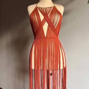 May include: A rust-colored halter top with a gold chain detail and long fringe. The top is made of a woven material and has a criss-cross design.