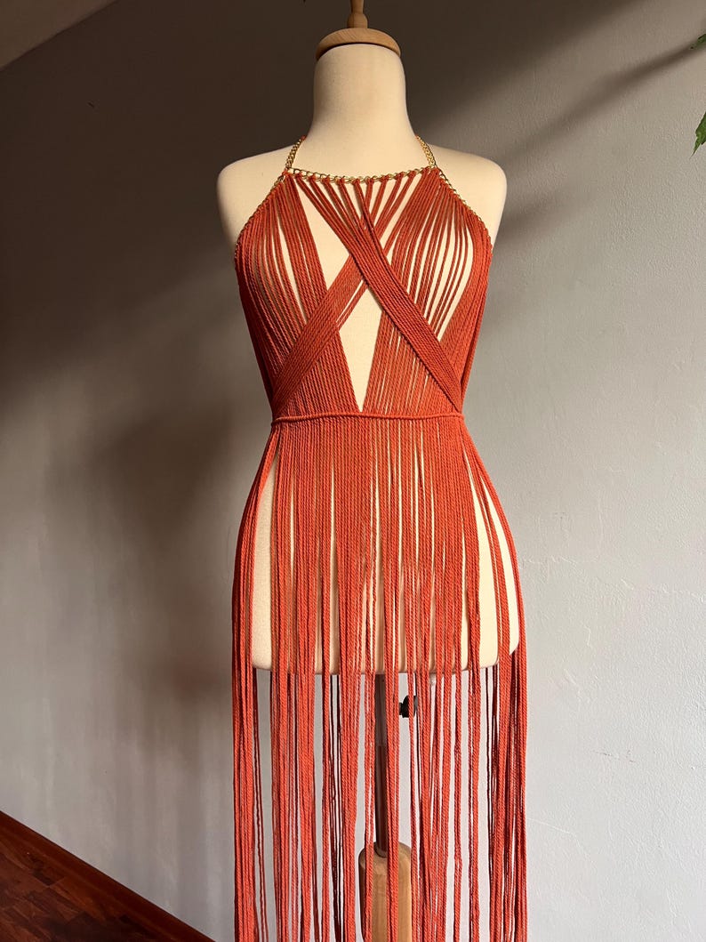 May include: A rust-colored halter top with a gold chain detail and long fringe. The top is made of a woven material and has a criss-cross design.