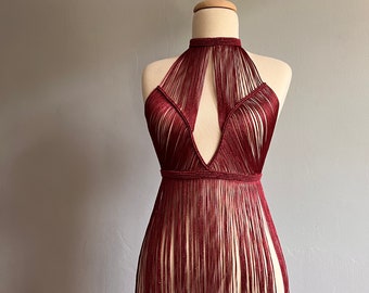 Puede incluir: Un top halter de color granate con detalles de flecos. El top está hecho de un material fino y en forma de cuerda y tiene un escote en V profundo.