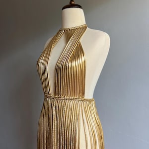 Puede incluir: Vestido dorado con cuello halter, escote pronunciado y laterales abiertos. El vestido est&aacute; hecho de hebras met&aacute;licas verticales, creando un efecto de flecos. El vestido se exhibe en un maniqu&iacute; sobre un fondo gris.