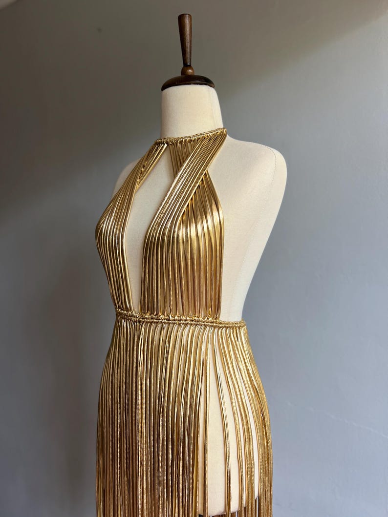 Puede incluir: Vestido dorado con cuello halter, escote pronunciado y laterales abiertos. El vestido est&aacute; hecho de hebras met&aacute;licas verticales, creando un efecto de flecos. El vestido se exhibe en un maniqu&iacute; sobre un fondo gris.