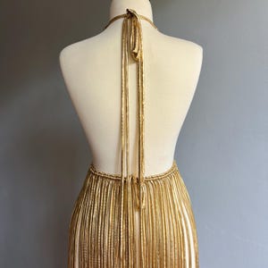 Puede incluir: Vestido con flecos dorados sobre un maniqu&iacute;. El vestido presenta un cuello halter y una falda con flecos. Los flecos dorados cuelgan verticalmente, creando un efecto fluido. El vestido se exhibe sobre un fondo gris neutro.