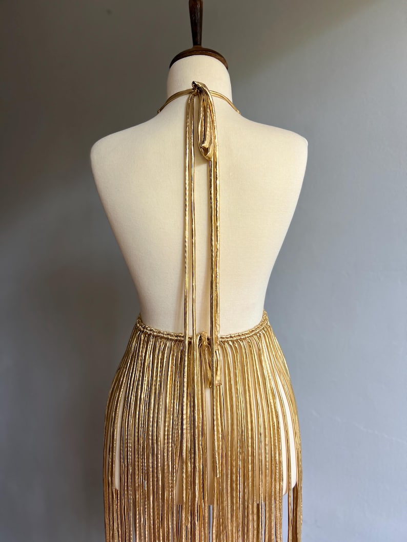 Puede incluir: Vestido con flecos dorados sobre un maniqu&iacute;. El vestido presenta un cuello halter y una falda con flecos. Los flecos dorados cuelgan verticalmente, creando un efecto fluido. El vestido se exhibe sobre un fondo gris neutro.