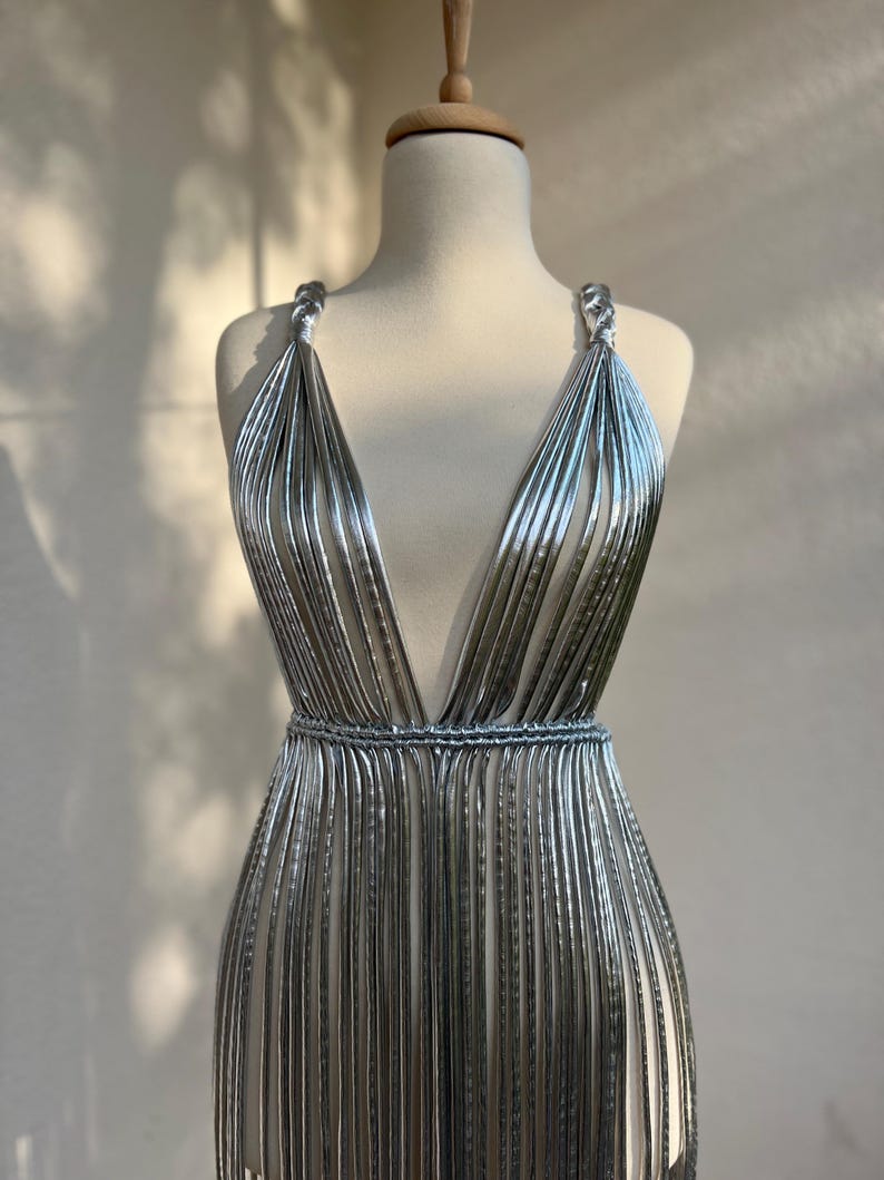 May include: A silver, sleeveless dress with a plunging neckline and a cinched waist. The dress is made of vertical, metallic strips that create a flowing, textured effect. The straps are twisted, and the dress is displayed on a mannequin.