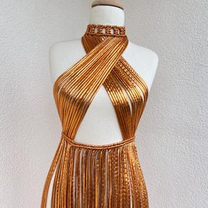 Op de afbeelding: Een goudkleurige, handgemaakte jurk, tentoongesteld op een mannequin. De jurk heeft een halterhalslijn en een gekruist lijfje. De rok heeft een franje-achtig detail. De jurk is gemaakt van een glanzend, metallic materiaal.