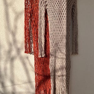 May include: Two long, crocheted dresses hanging on hangers. The dress on the left is a bright orange color, and the dress on the right is a light beige color. Both dresses have a loose, open weave and fringe detailing.