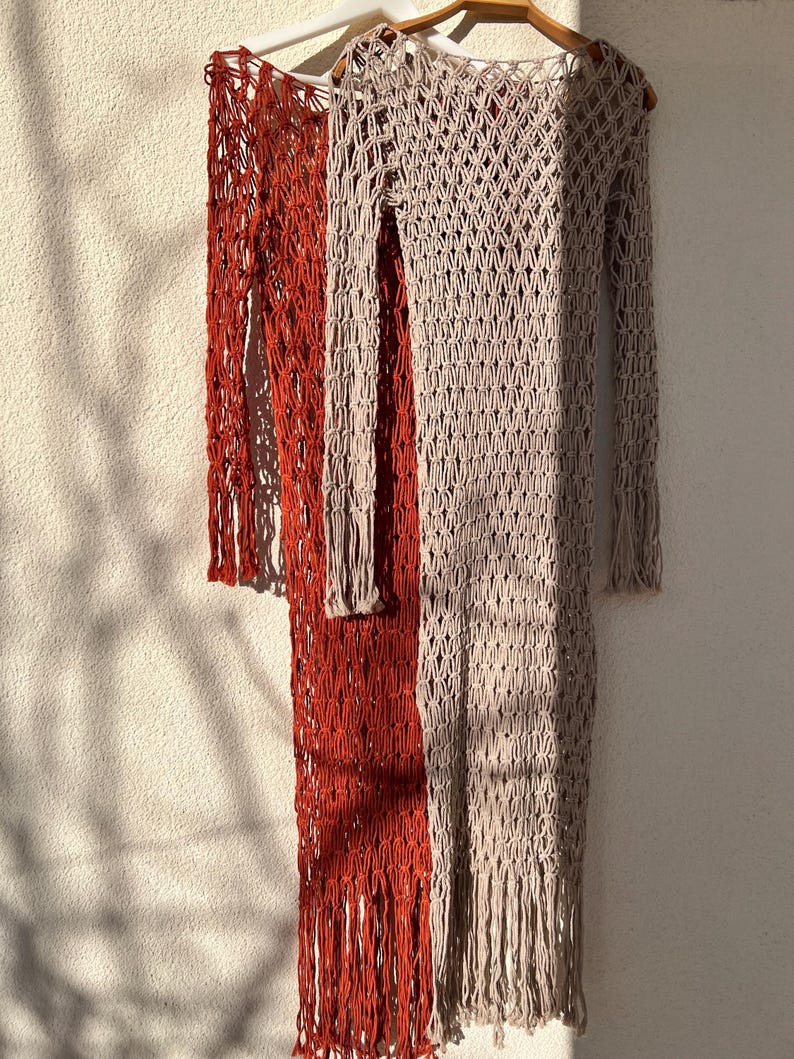 May include: Two long, crocheted dresses hanging on hangers. The dress on the left is a bright orange color, and the dress on the right is a light beige color. Both dresses have a loose, open weave and fringe detailing.